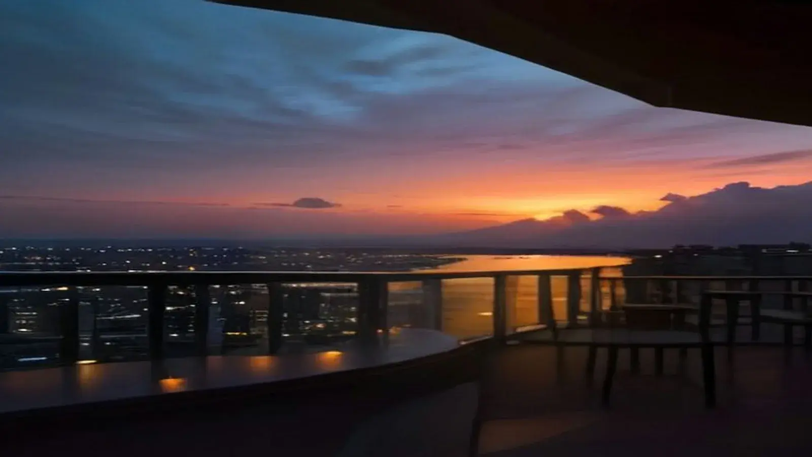 Illustrative scene: the view from a high rooftop terrace over Pattaya Bay at sunset, with an orange and violet sky, rain clouds building over the hills, city lights coming on below the glass railing and empty lounge seats on the deck.