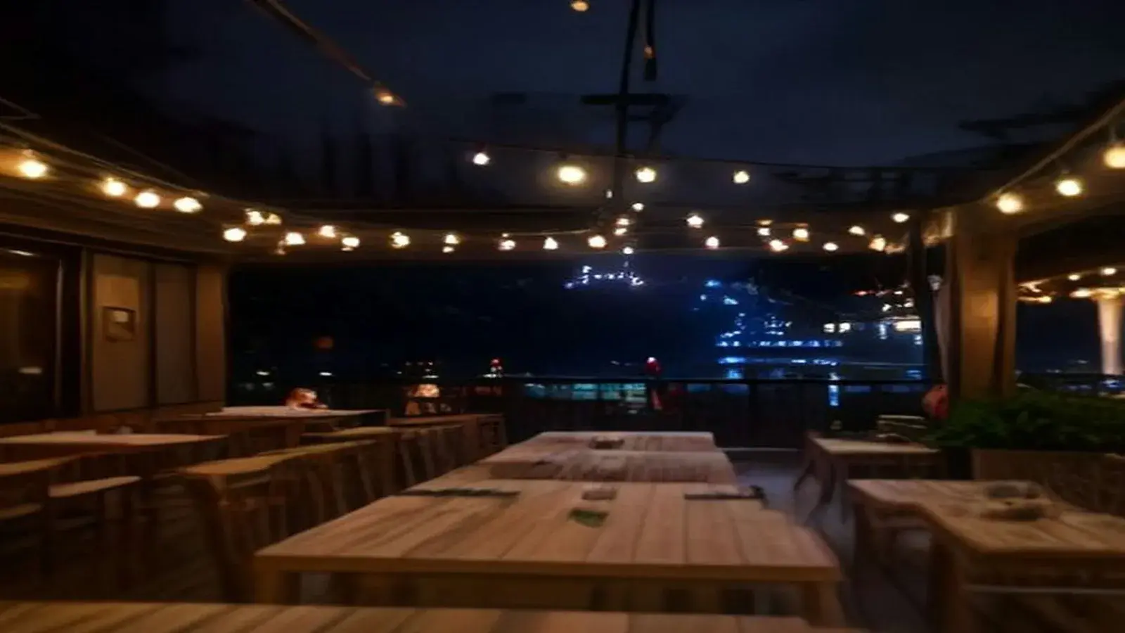Illustrative scene: long wooden tables under a covered open-air beer garden roof at night in Pattaya, with warm string lights overhead and the lights of the bay glowing beyond the railing, the kind of sheltered seating suited to a rainy Sunday night.