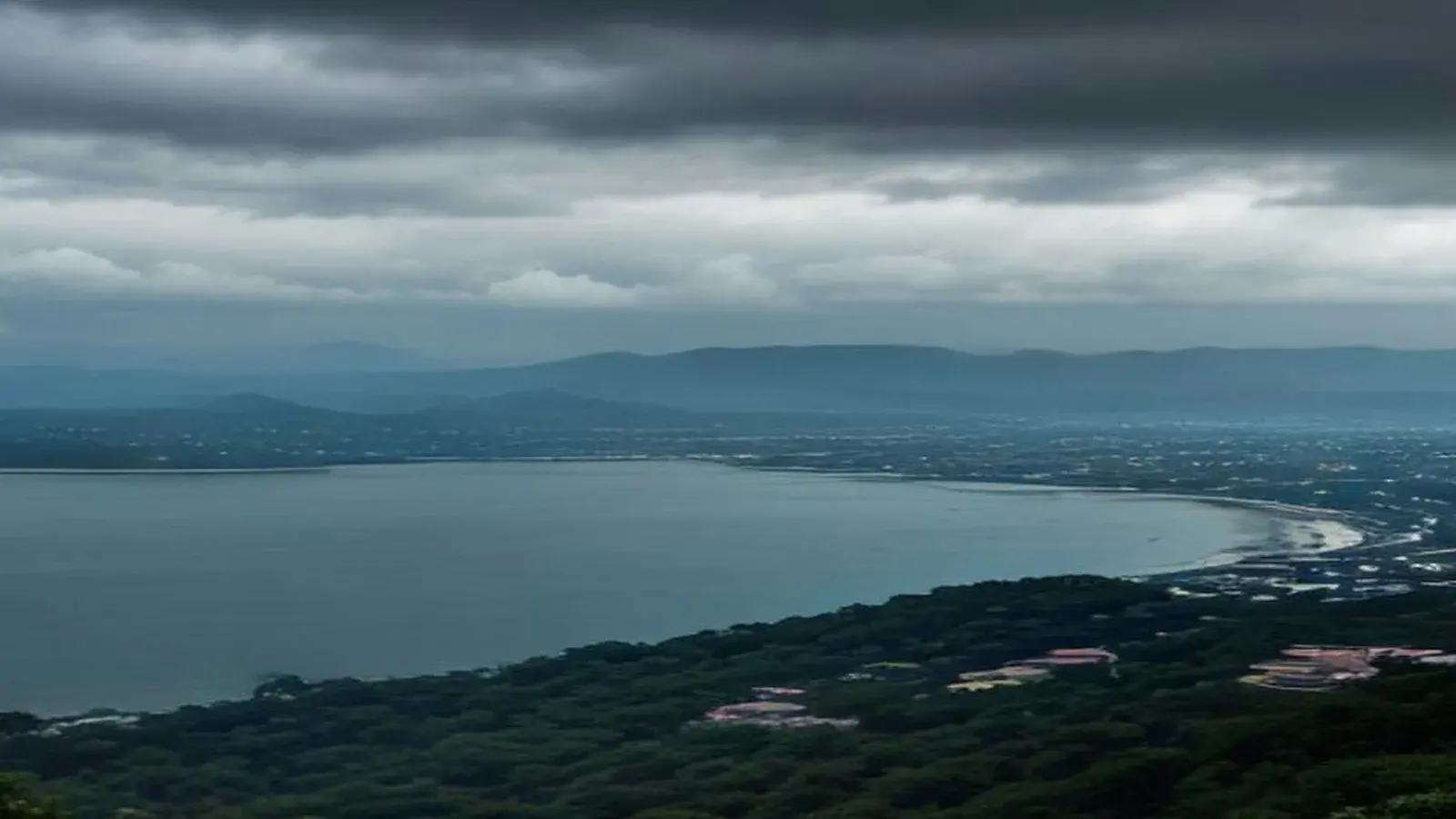 Illustrative scene: heavy grey rain clouds sit low over Pattaya Bay seen from the wooded Pratamnak hill, with the curve of the beach and the city below and the hills behind fading into haze, matching today's TMD heavy rain warning for the East.